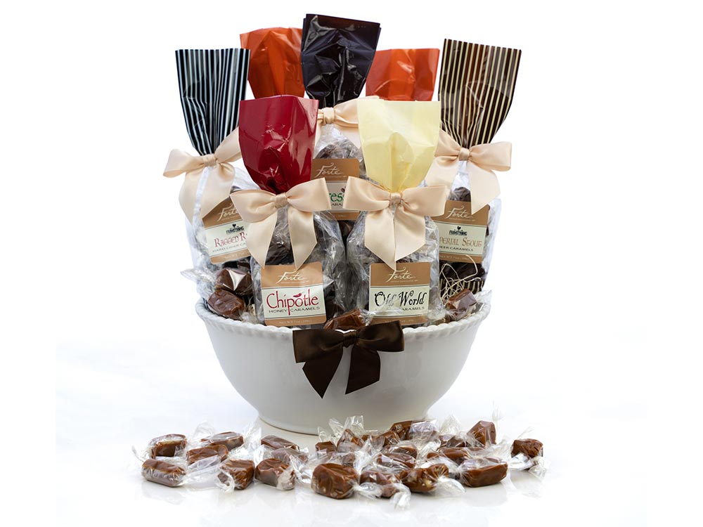 White bowl filled with 7 beribboned bags of assorted caramels and wrapped caramels on the table in the foreground