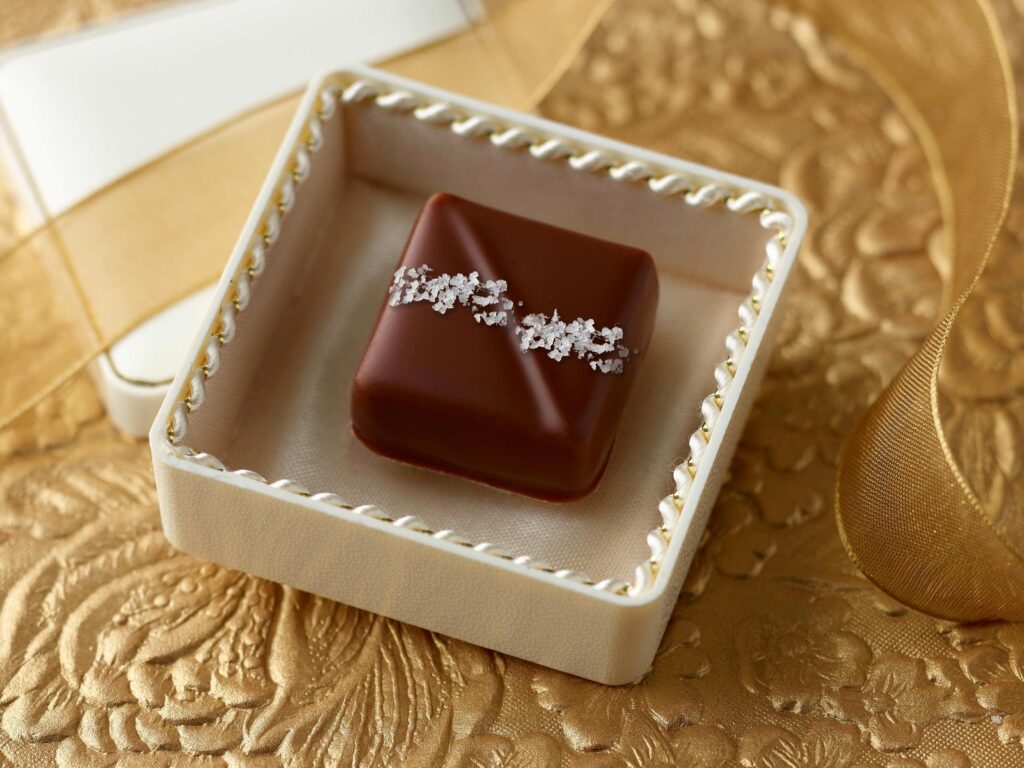 Sea salt milk chocolate caramel artfully placed in a small jewel box with a loose gold ribbon on a gold surface