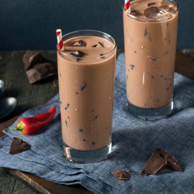 2 glasses of iced aztec cocoa with red and white stripped straws on blue cloths with chocolate shavings a large red pepper