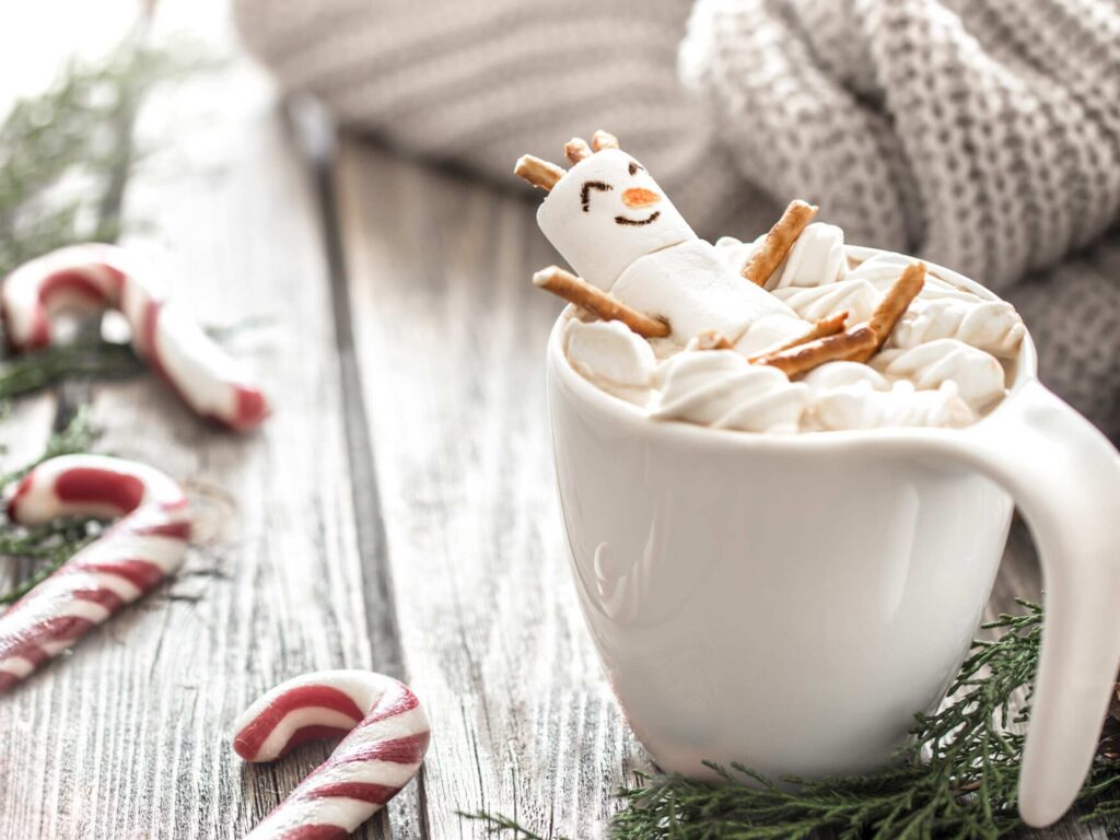 Marshmallow snowman with pretzel arms floating in a white mug of peppermint hot cocoa with 3 peppermint sticks and green foliage on a grey brown background