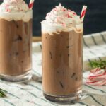 2 glasses filled with iced peppermint cocoa topped with whipped cream sprinkled with peppermint shavings and red and white stripped straws placed on a blue and white striped cloth with peppermint sticks and rosemary twigs in background
