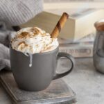 An overflowing mug of vanilla hot cocoa in a grey cup with whipped cream and a stripped rolled wafer cookie with books and a sweater in the background