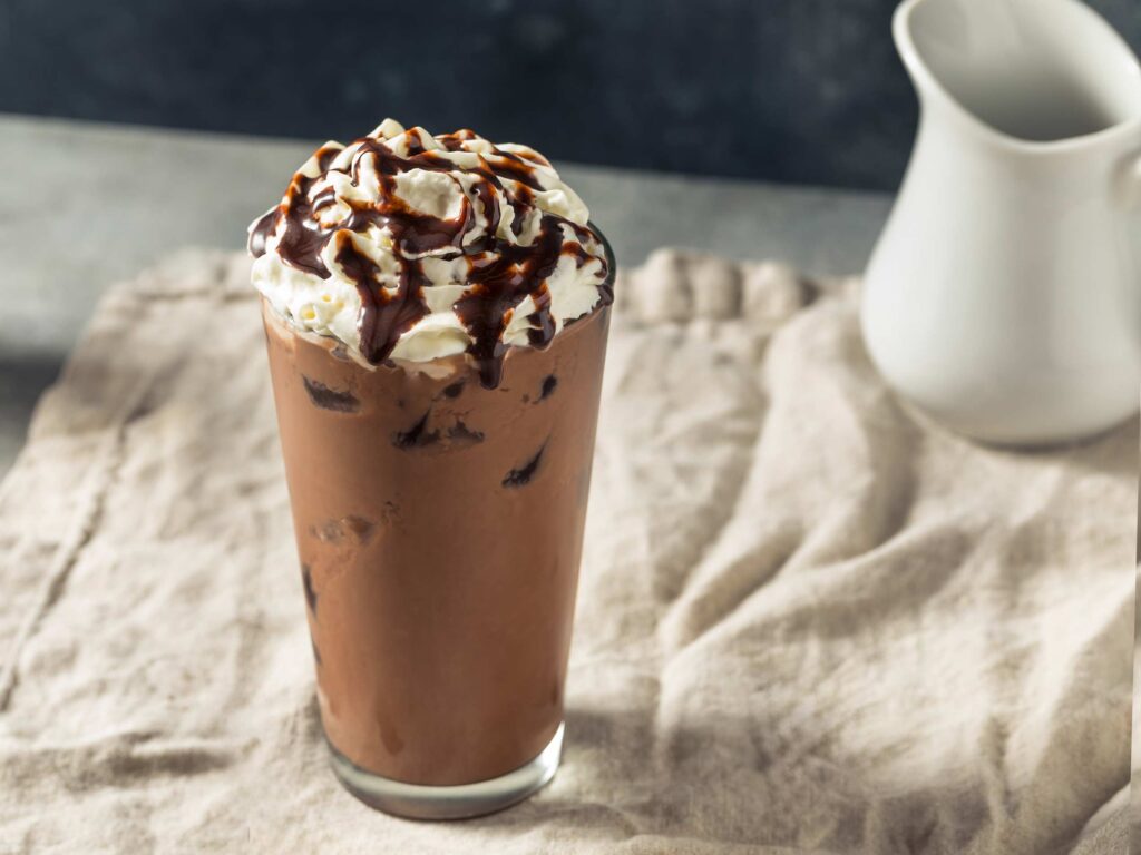 Delicious iced vanilla cocoa in a tall glass with whipped cream and chocolate drizzles on top of a white linen cloth with a cream pitcher in the background