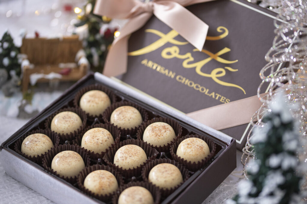 12 piece box of winter seasonal eggnog truffles in brown box with pink ribbon surrounded with festive holiday decorations. Artisan eggnog truffles for Christmas. Limited Edition Holiday Truffles Forte Chocolate