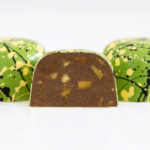 Trio of pistachio truffles in bright green with black and yellow splatters with center piece halved showing chunks of pistachio in creamy milk chocolate ganache