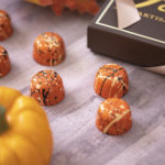 8 pumpkin spice truffles in orange with black and cream colored splatters arranged on a wooden table with small pumpkin in foreground and brown Forte box in background