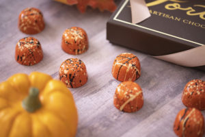 8 pumpkin spice truffles in orange with black and cream colored splatters arranged on a wooden table with small pumpkin in foreground and brown Forte box in background. Seasonal chocolate flavors, artisan holiday chocolate