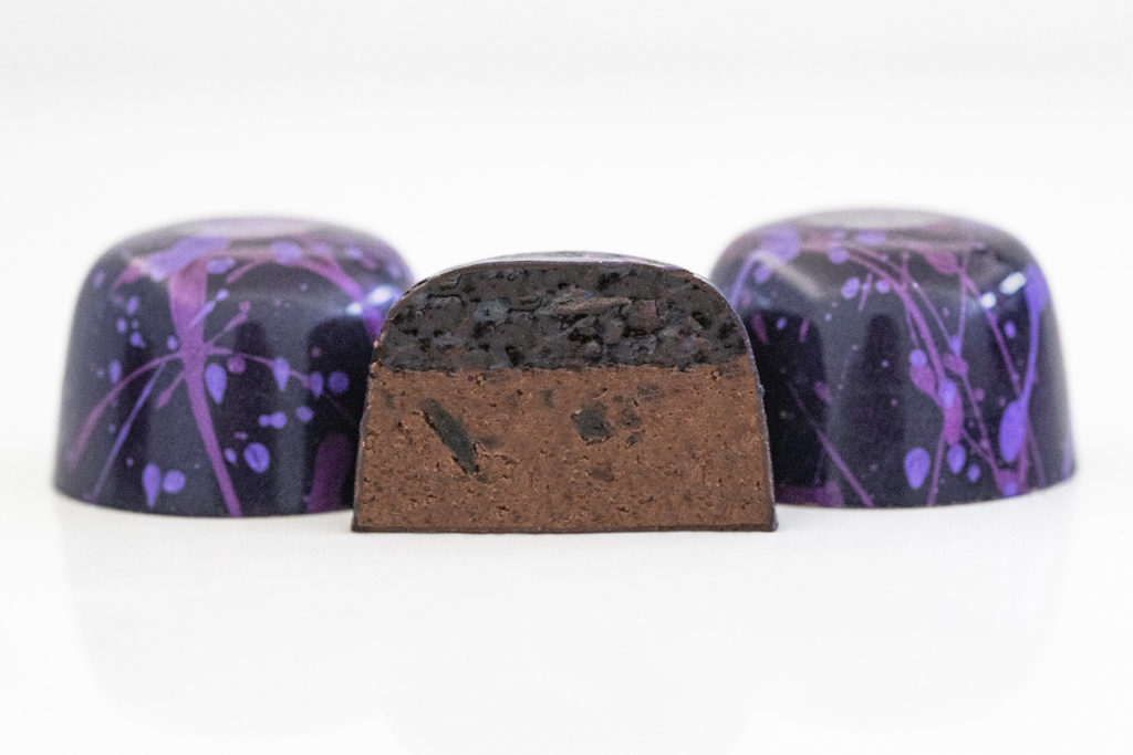 3 raspberry fig truffles in deep purple with light purple and pink splatters with center truffle halved to show chocolate ganache and a dollop of raspberry fig preserves