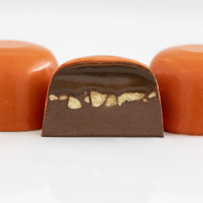 3 pecan turtle truffles in deep orange with center truffle halved showing pecan and chocolate ganache with a dollop of creamy caramel on top