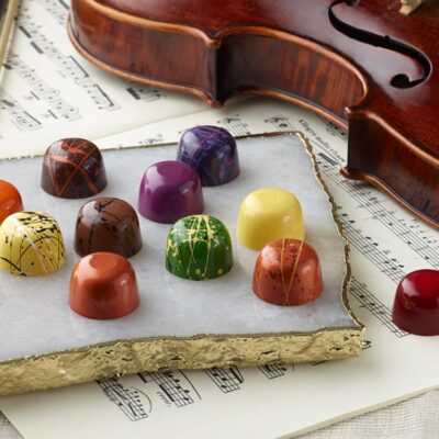 12 Signature assorted chocolate truffles artfully arranged on a white marble slab with 1 red truffle on top of violin music next to a violin with bow