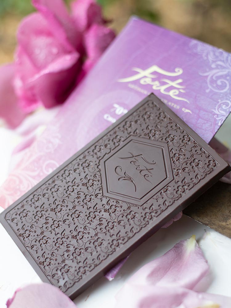 Unwrapped Forte 70% chocolate bar with logo and chocolate mold design on top of purple packaging in front of a purple flower and petals
