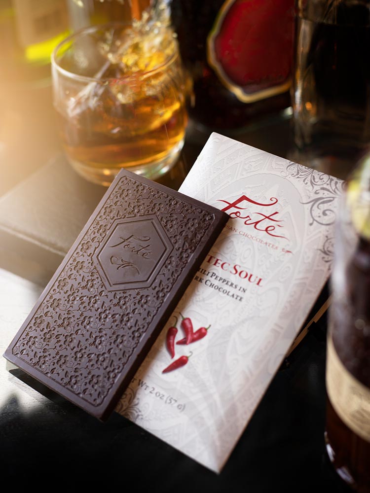 Unwrapped Forte Aztec Soul chocolate bar showing Forte logo and bar mold design atop the red and white packaging with a cocktail glass filled with ice and amber liquid in background
