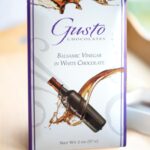 Gusto balsamic vinegar in white chocolate bar with purple edges on light wood tray with ramekin of balsamic vinegar
