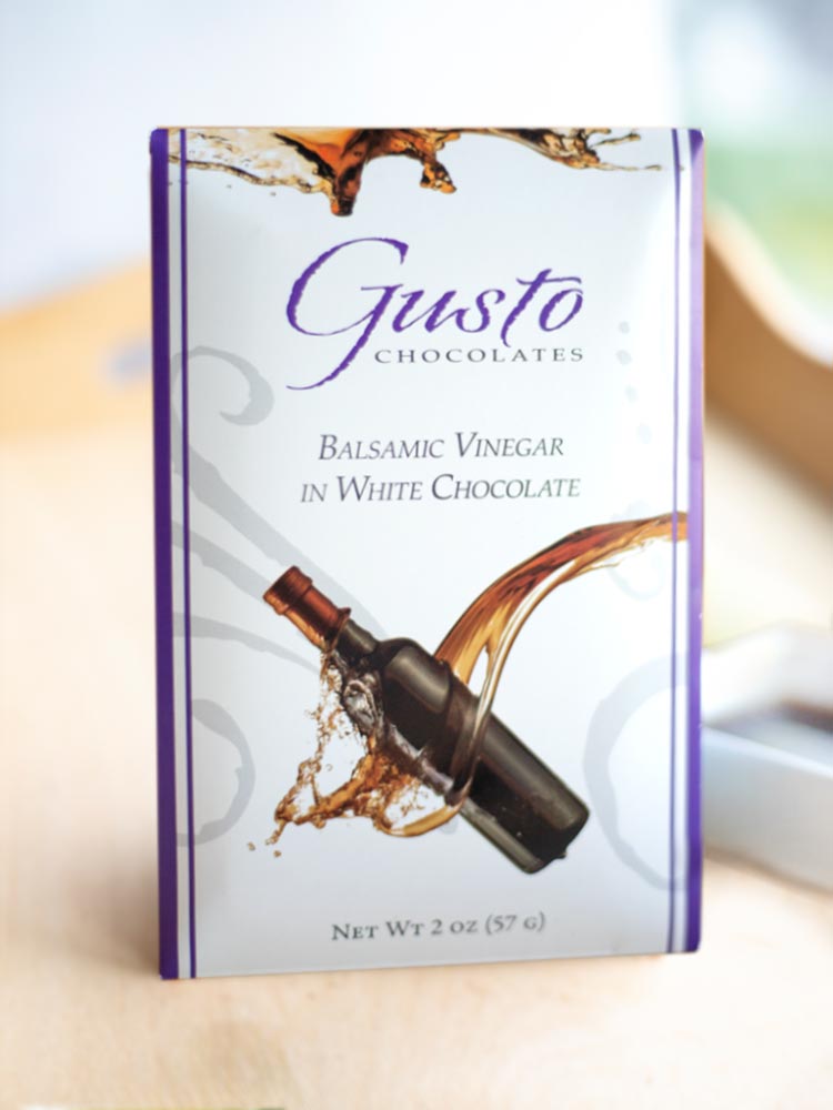 Gusto balsamic vinegar in white chocolate bar with purple edges on light wood tray with ramekin of balsamic vinegar