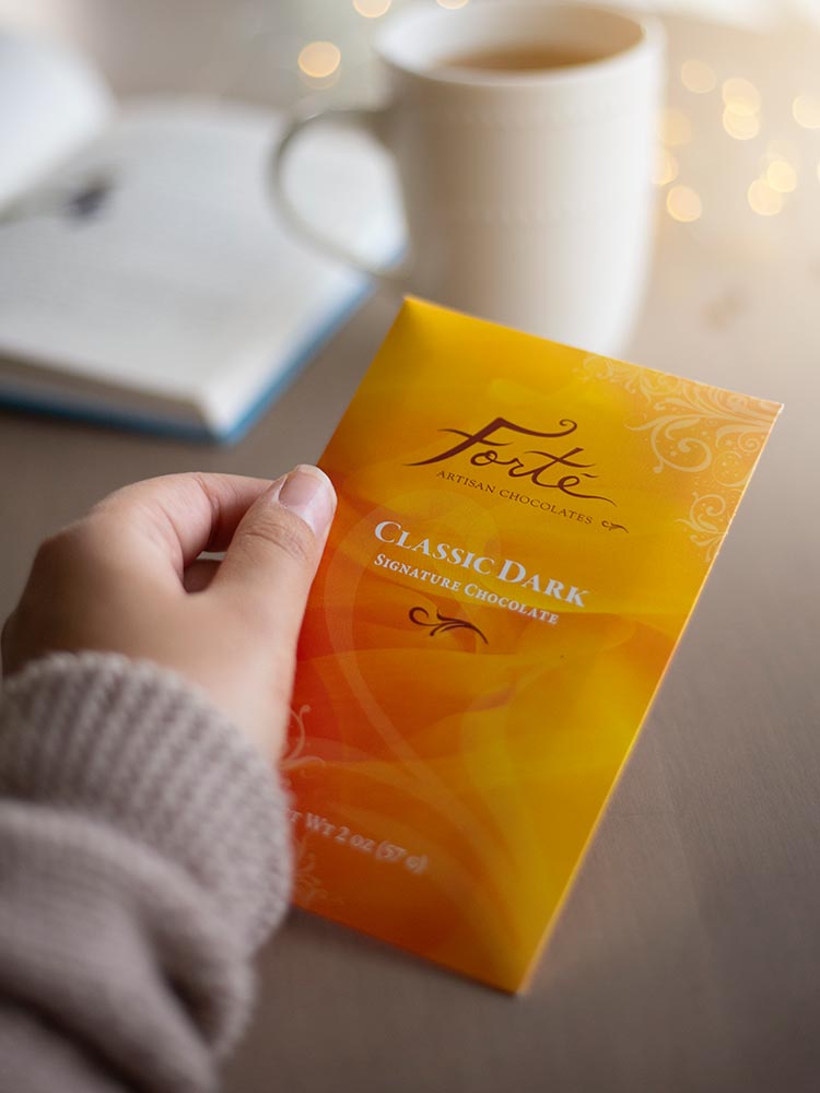 Classic dark chocolate bar package held in a hand with open book and cup of tea in background