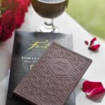 An unwrapped Forte Fortunato bar showing the Forte logo and bar mold design of cocoa flowers sits atop the packaging for the bar Red roses and petals surround the bar with a hearty glass of red wine in the top of the frame