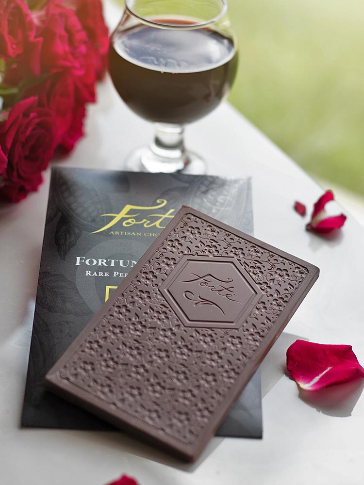 An unwrapped Forte Fortunato bar showing the Forte logo and bar mold design of cocoa flowers sits atop the packaging for the bar Red roses and petals surround the bar with a hearty glass of red wine in the top of the frame