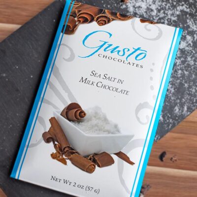 Gusto sea salt in milk chocolate bar package on slate board dusted with sea salt and chocolate shavings
