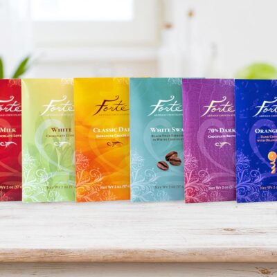 6 bars from Forte collection lined up on a shelf with small chocolate bites in foreground and greenery in back