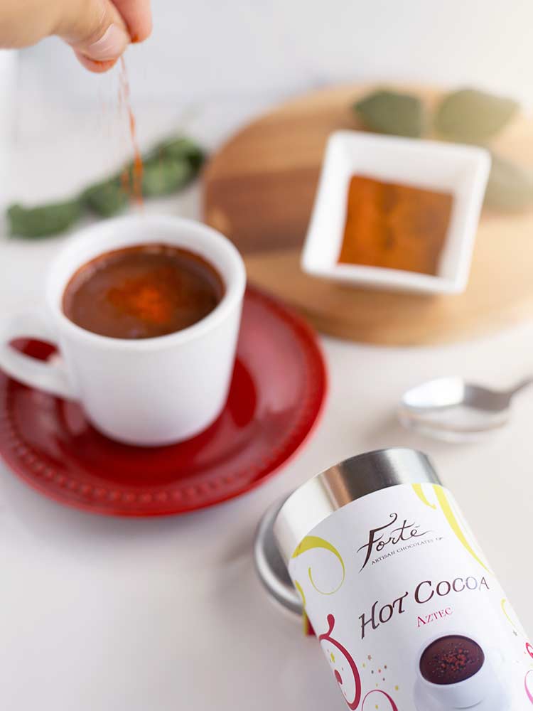 White mug on a red saucer of aztec hot cocoa with a hand sprinkling red chili powder on top A tin of Forte Aztec hot cocoa lies on its side with a ramekin of chili powder in the background