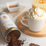 White mug of salted caramel hot cocoa with whipped cream and active drizzle of caramel next to a tipped over cocoa tin and spoon with caramel containers in background