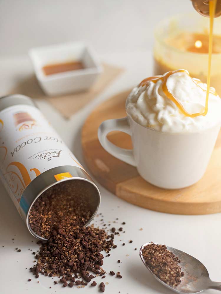White mug of salted caramel hot cocoa with whipped cream and active drizzle of caramel next to a tipped over cocoa tin and spoon with caramel containers in background