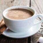 White mug in saucer with vanilla hot cocoa with two vanilla stalks on saucer and 4 small marshmallows in foreground