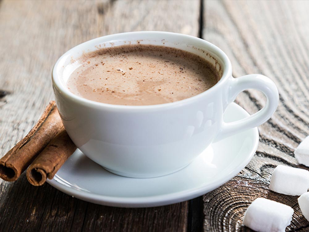 White mug in saucer with vanilla hot cocoa with two vanilla stalks on saucer and 4 small marshmallows in foreground