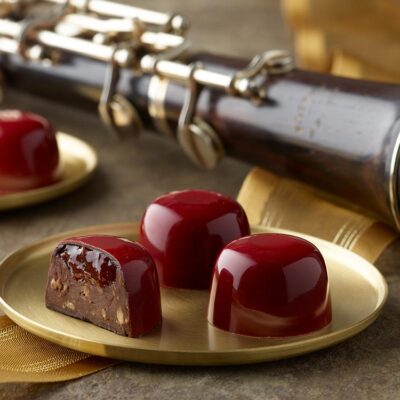 5 bright red cherry almond truffles arranged on two small gold plates with a gold ribbon and oboe in the background One truffle is sliced in half showing a layer of cherry preserves atop an almond and chocolate ganache