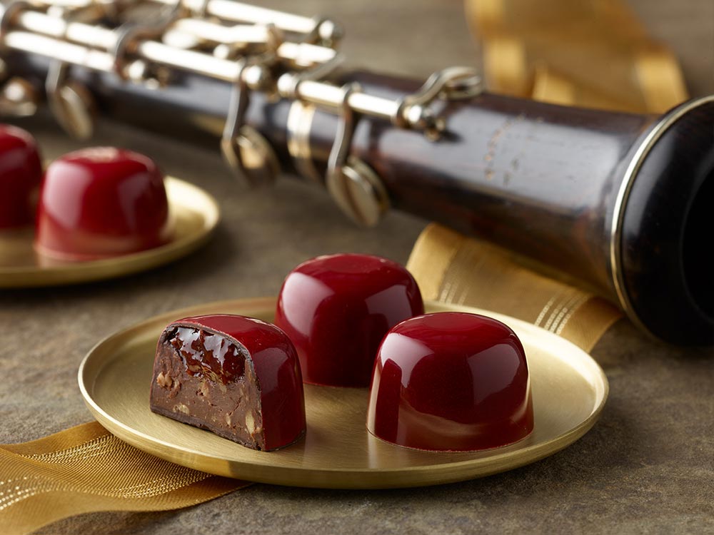 5 bright red cherry almond truffles arranged on two small gold plates with a gold ribbon and oboe in the background One truffle is sliced in half showing a layer of cherry preserves atop an almond and chocolate ganache