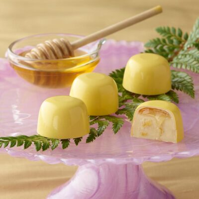 4 honey lemon truffles atop a bright pink cake stand with a fern leaf and small honey bowl with a honey dipper One truffle is sliced in half showing a layer of honey atop a lemony ganache inside a white chocolate cup in bright yellow