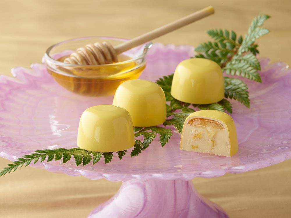 4 honey lemon truffles atop a bright pink cake stand with a fern leaf and small honey bowl with a honey dipper One truffle is sliced in half showing a layer of honey atop a lemony ganache inside a white chocolate cup in bright yellow
