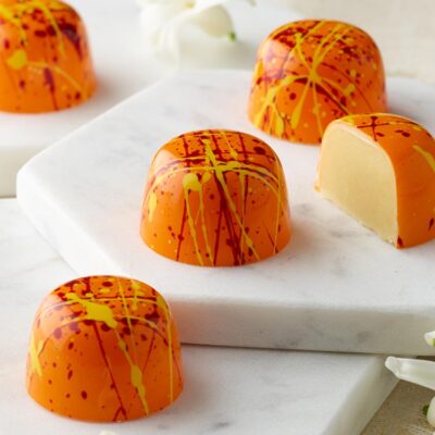 5 bright passion fruit truffles with red and yellow splatters on an orange cup placed on white marble with flowers in background