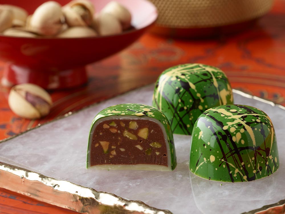 3 pistachio truffles in bright green with yellow and black splatters on a marble square with pistachio nuts in a red bowl in background One truffle is sliced open showing large pistachio chunks in the milk chocolate ganache and a white chocolate cup
