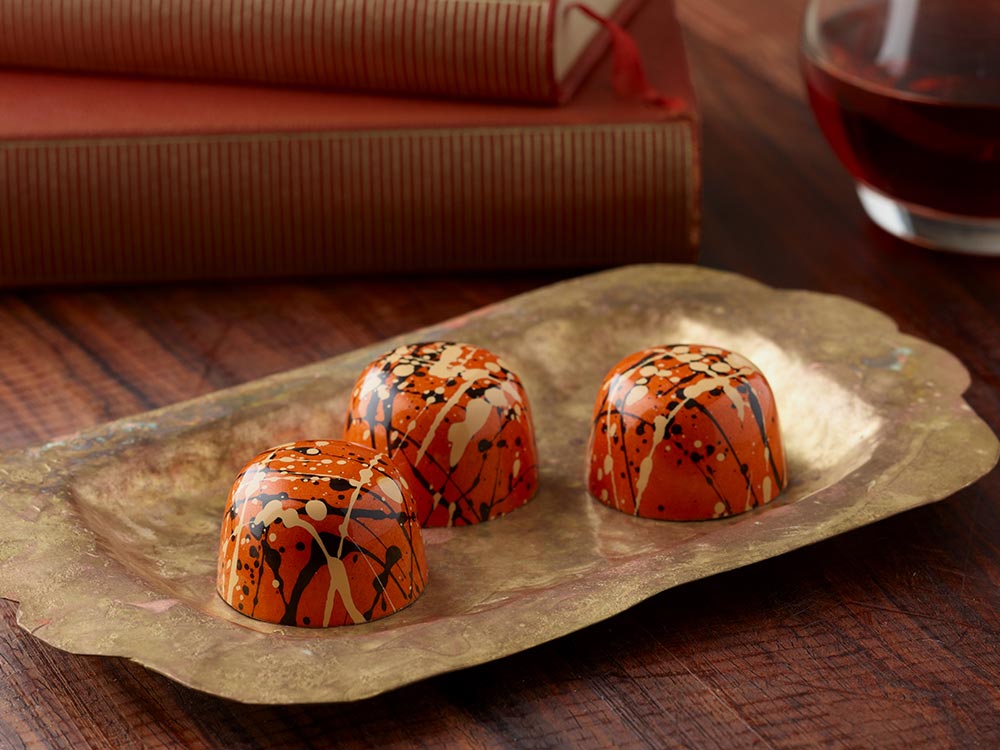 3 pumpkin spice truffles in orange with tan and dark brown splatters artfully arranged on an oblong bronze platter in front of two red books and a glass of something tasty