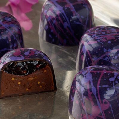 Close-up of 5 raspberry fig truffles arranged on a silver platter with two pink orchids and a crystal glass in background One truffle is sliced in half showing the layer of raspberry preserves atop the fig and milk chocolate ganache inside a dark chocolate cup splattered with pink and purple