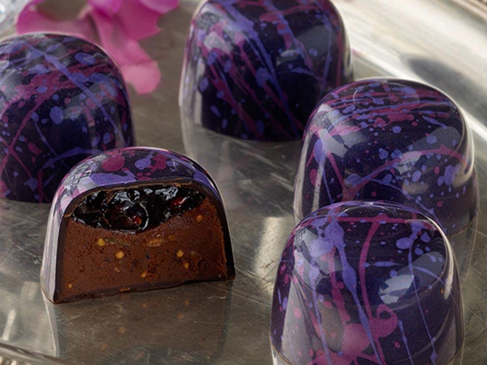 Close-up of 5 raspberry fig truffles arranged on a silver platter with two pink orchids and a crystal glass in background One truffle is sliced in half showing the layer of raspberry preserves atop the fig and milk chocolate ganache inside a dark chocolate cup splattered with pink and purple