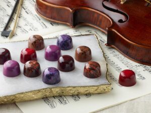 12 assorted chocolate truffles artfully arranged on a white marble slab with 1 red truffle on top of violin music next to a violin with bow