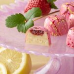5 strawberry lemonade truffles in bright pink with red and yellow splatters on top of a pink cake plate with mint and large strawberry above a sliced lemon One truffle is sliced showing a splash of strawberry preserves atop the lemonade ganache in a white chocolate shell