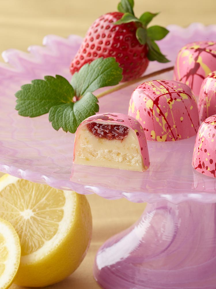 5 strawberry lemonade truffles in bright pink with red and yellow splatters on top of a pink cake plate with mint and large strawberry above a sliced lemon One truffle is sliced showing a splash of strawberry preserves atop the lemonade ganache in a white chocolate shell