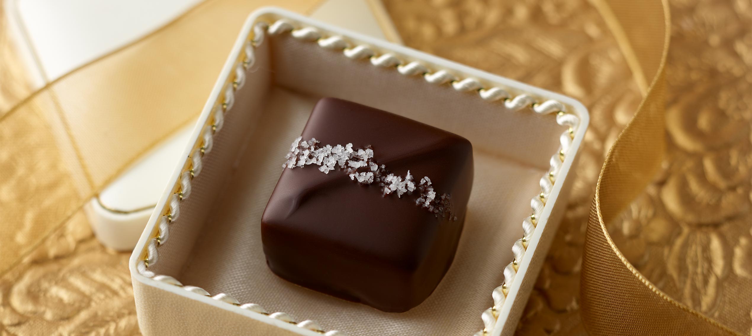 Dark chocolate caramel with band of sea salt placed in an ivory ring box with scalloped edge on top of a field of gold and gold ribbon