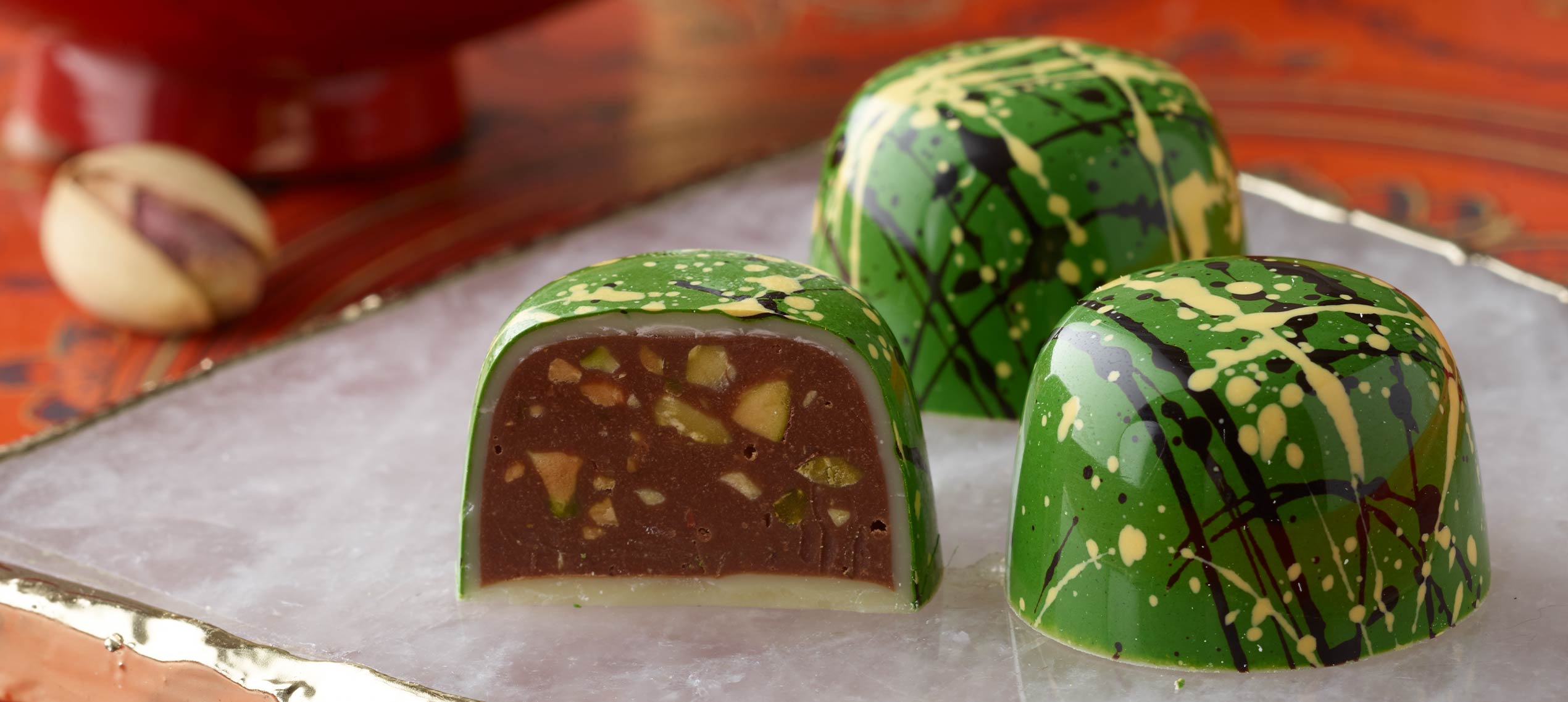 Pistachio truffle - two green with black and yellow splatters and one sliced in half - placed on a white tile with a pistachio in shell background