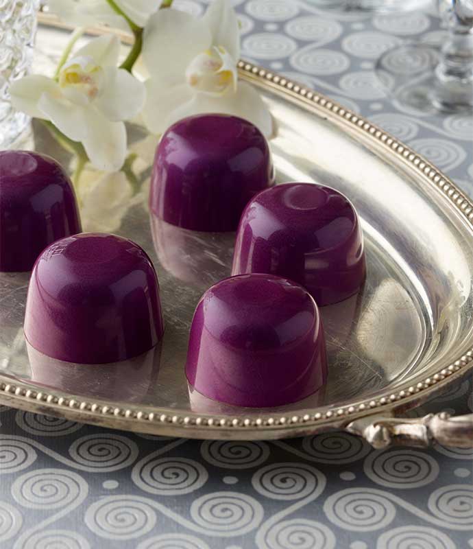 Five bright purple vegan superfood truffles on a silver platter with two orchids on a grey swirled background