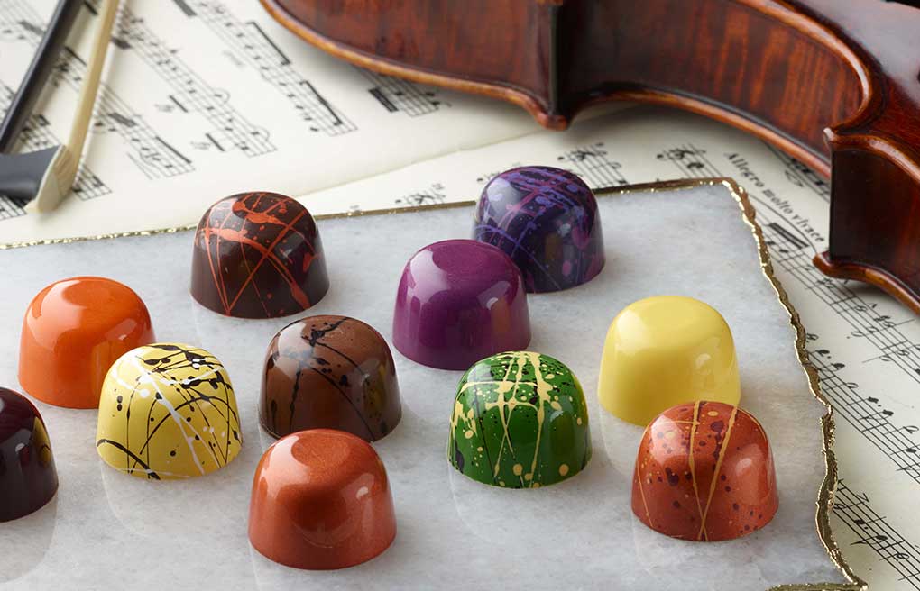 Assorted truffles in various colors on a white tile with gold edge on sheet music with violin and bow in background