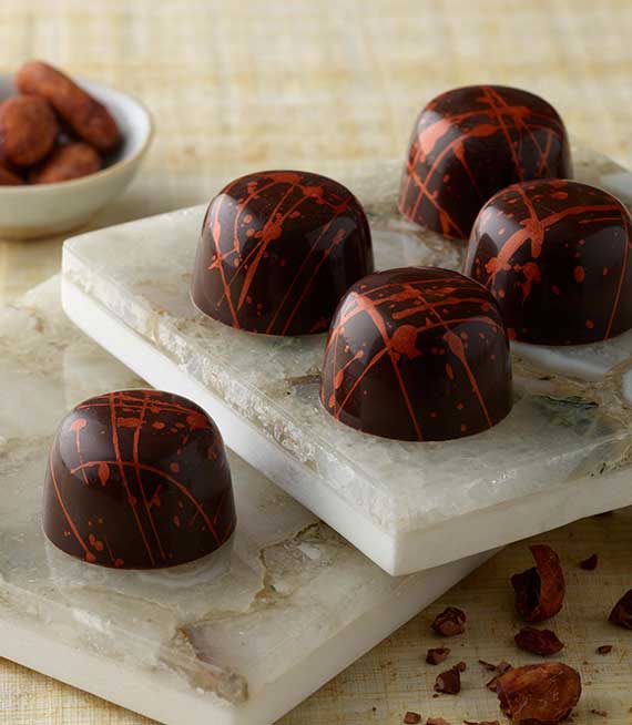 Five classic dark truffles with red-gold splatters placed on two white tiles with cocoa beans in background