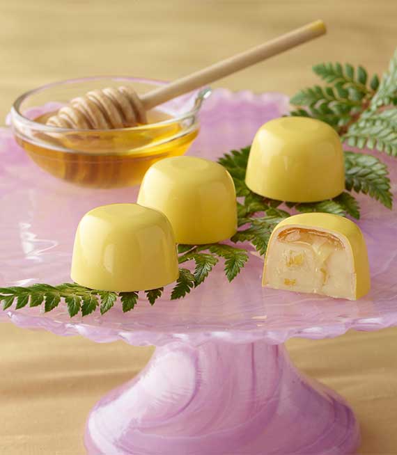 Three bright yellow honey lemon truffles with a fourth sliced in half showing lemon ganache and dripping honey on a pink cake stand with a ramekin of honey with a wooden honey dipper